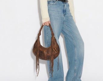 Puede incluir: Un suéter de cuello alto color crema de manga larga, pantalones vaqueros azul claro con cinturón negro y un bolso hobo de cuero marrón oscuro con asa trenzada y flecos largos, combinado con zapatos de cuero marrón oscuro.