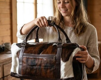 Puede incluir: Un bolso de viaje hecho de piel de vaca marrón y blanca con ribetes de cuero marrón oscuro, dos asas superiores resistentes, un bolsillo exterior horizontal con cremallera y herrajes metálicos. El bolso tiene una forma rectangular.
