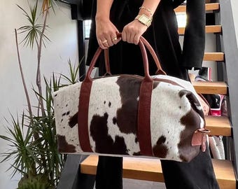Puede incluir: Una bolsa de lona rectangular grande con un estampado de piel de vaca en blanco y marrón. La bolsa tiene asas y detalles de cuero marrón. La persona está de pie en escaleras de madera.