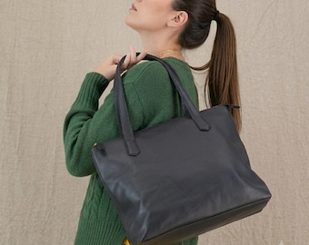 Puede incluir: Un bolso tote de cuero negro liso con dos correas para el hombro, que presenta una forma rectangular y un acabado suave. El bolso se sostiene por el costado, mostrando sus líneas limpias, diseño minimalista y construcción funcional.