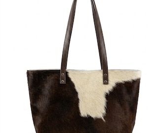 Puede incluir: Un bolso tote rectangular con un estampado de piel de vaca en tonos marrón y crema. El bolso tiene dos asas de cuero marrón oscuro y está diseñado para transportar objetos. El bolso está hecho de un material suave y texturizado.