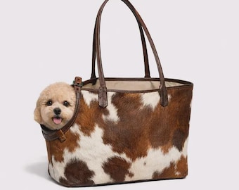 Puede incluir: Un bolso tote de piel de vaca marrón y blanco con un perro pequeño asomando. El bolso tiene asas y ribetes de cuero marrón. El perro es de color marrón claro y mira por una abertura circular en el lateral del bolso.