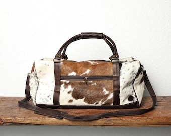 Puede incluir: Un bolso de viaje confeccionado en piel de vaca con manchas marrones y blancas. El bolso cuenta con ribetes de cuero marrón oscuro, dos asas superiores, un bolsillo frontal con cremallera y una correa de hombro extraíble con sección acolchada.