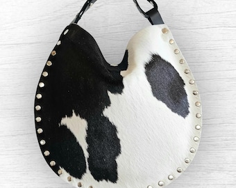 Puede incluir: Un bolso hobo de piel de vaca en blanco y negro con una correa de cuero negro. El bolso tiene una forma redondeada con detalles de tachuelas plateadas en los bordes. El diseño del bolso muestra un patrón natural de manchas negras sobre fondo blanco.