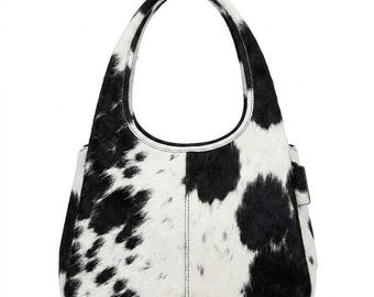 Puede incluir: Un bolso de mano de piel de vaca con un estampado de manchas blancas y negras. El bolso tiene forma redondeada con un asa y un pequeño bolsillo lateral. El bolso está hecho de un material suave y texturizado.