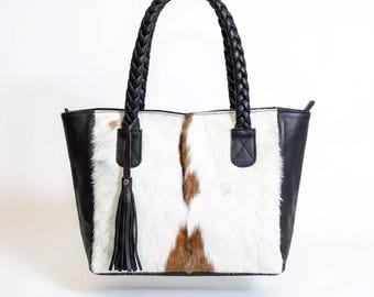 Puede incluir: Un bolso tote con un panel frontal de piel de vaca con pelo blanco, marrón y negro. Los laterales y la base son de cuero negro. Tiene dos asas trenzadas de cuero negro y una borla negra.