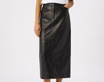 Puede incluir: Un conjunto femenino con una camiseta blanca y una falda midi de cuero negro. La falda tiene cintura alta y corte recto. El conjunto se completa con sandalias negras.