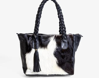 Puede incluir: Bolso tote de piel de vaca en blanco y negro con asas de cuero trenzado. El bolso tiene un ribete de cuero negro y una borla de cuero negro. Cuenta con un cierre de cremallera y está diseñado para transportar objetos.