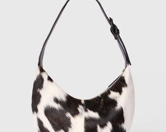 Puede incluir: Un bolso en forma de media luna con un estampado de piel de vaca en blanco y negro. El bolso tiene una correa de cuero negro y una silueta curva. El bolso está diseñado para llevarse al hombro.