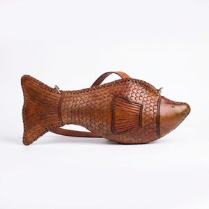 May include: A brown leather handbag shaped like a fish, featuring embossed scale patterns, a stitched fin detail, a pointed tail, and a thin adjustable shoulder strap attached with small metal hardware rings on the top of the body.