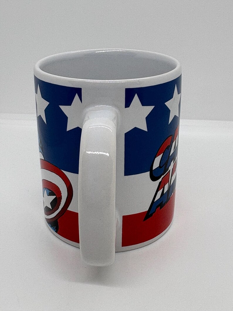 Puede incluir: Taza de cer&aacute;mica blanca con un dise&ntilde;o inspirado en el Capit&aacute;n Am&eacute;rica. La taza tiene un asa blanca y un dise&ntilde;o con colores azul, rojo y blanco. El dise&ntilde;o incluye estrellas, rayas y las palabras "Captain America".