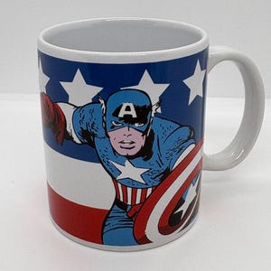 Puede incluir: Taza de cer&aacute;mica blanca con el Capit&aacute;n Am&eacute;rica al estilo c&oacute;mic. La taza tiene un fondo azul con estrellas blancas y rayas rojas y blancas. El asa es blanca.