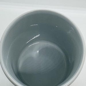 Puede incluir: Una taza de cer&aacute;mica blanca con interior gris claro. La taza tiene forma redondeada y un asa, y est&aacute; sobre un fondo blanco. El interior de la taza es liso y brillante.