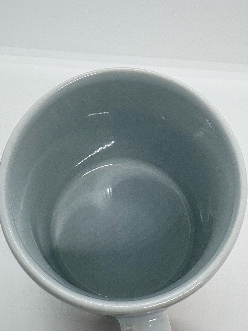 Puede incluir: Una taza de cer&aacute;mica blanca con interior gris claro. La taza tiene forma redondeada y un asa, y est&aacute; sobre un fondo blanco. El interior de la taza es liso y brillante.