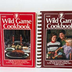 May include: Two spiral-bound cookbooks titled "Wild Game Cookbook" by NAHC. The covers are red and maroon, featuring images of food and people. The red cover has a table setting with food, and the maroon cover shows a family. The books are for recipes submitted by the North American Hunting Club.