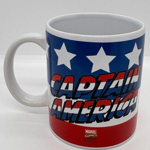 Puede incluir: Taza de cer&aacute;mica blanca con las palabras "CAPTAIN AMERICA" en rojo y azul, sobre un fondo azul con estrellas blancas. La taza tiene un asa blanca y el logotipo de Marvel Comics.