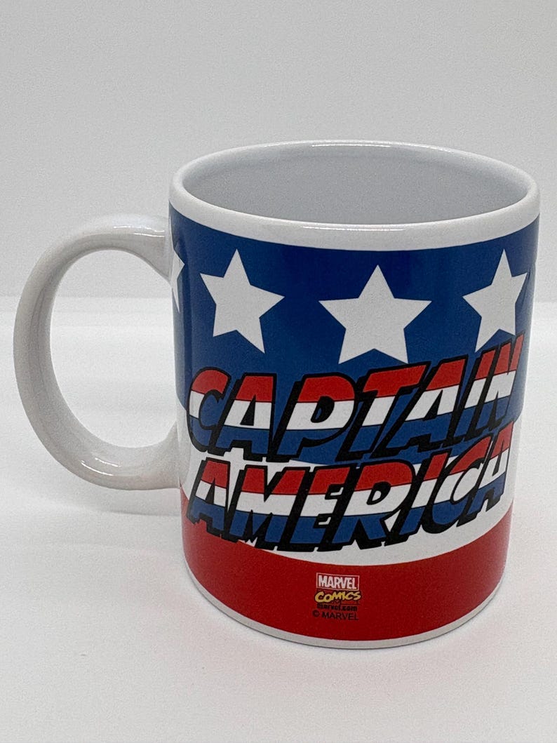 Puede incluir: Taza de cer&aacute;mica blanca con las palabras "CAPTAIN AMERICA" en rojo y azul, sobre un fondo azul con estrellas blancas. La taza tiene un asa blanca y el logotipo de Marvel Comics.