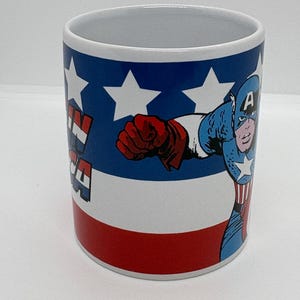 Puede incluir: Taza de cer&aacute;mica blanca con el Capit&aacute;n Am&eacute;rica en acci&oacute;n. La taza tiene un dise&ntilde;o rojo, blanco y azul con estrellas y las palabras "Captain America". El superh&eacute;roe est&aacute; representado con su traje cl&aacute;sico.