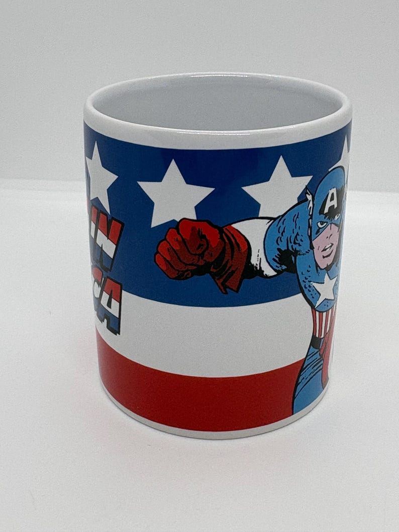 Puede incluir: Taza de cer&aacute;mica blanca con el Capit&aacute;n Am&eacute;rica en acci&oacute;n. La taza tiene un dise&ntilde;o rojo, blanco y azul con estrellas y las palabras "Captain America". El superh&eacute;roe est&aacute; representado con su traje cl&aacute;sico.