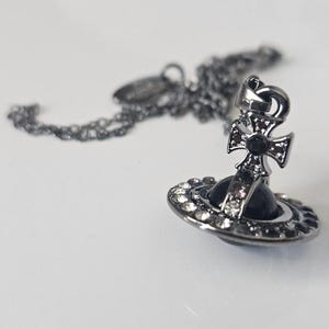 Puede incluir: Collar con colgante plateado con un colgante en forma de planeta. El colgante tiene un globo negro en el centro, rodeado de cristales transparentes. Una cruz está encima. La cadena también es plateada.