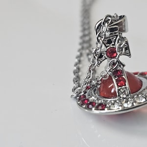 Peut inclure: Collier pendentif argenté avec un motif d'orb rouge, un cadre argenté et des ornements en cristaux rouges et transparents. Le pendentif est suspendu à une fine chaîne. Un accessoire de mode sur fond blanc.