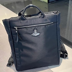 Puede incluir: Mochila negra de nailon con asa superior y correas ajustables para los hombros. Cuenta con un bolsillo frontal con cremallera y un emblema plateado con el logotipo. La bolsa tiene forma rectangular y un diseño elegante y moderno.