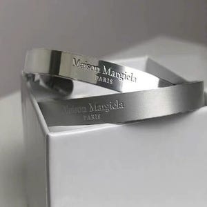 May include: Two silver-toned Maison Margiela Paris bracelets are displayed. The bracelets are stacked, with the brand name and location engraved on the surface. The bracelets are resting in a white box.