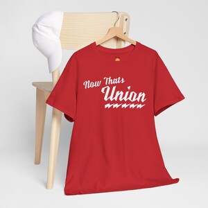 May include: A red t-shirt with the text "Now Thats Union" in white script. The shirt is hanging on a wooden hanger. A white baseball cap rests on a wooden chair in the background.