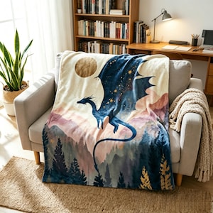 May include: A soft throw blanket featuring a dark blue dragon flying over a landscape of mountains and pine trees. The design includes a large golden moon and scattered stars against a cream-coloured background, with a watercolour-style aesthetic.
