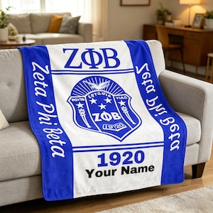 Op de afbeelding: Een blauw-witte Zeta Phi Beta deken op een bank. De deken toont de letters ZΦB, een wapen met Griekse tekst en het jaar 1920. De woorden "Zeta Phi Beta" staan verticaal op de zijkanten gedrukt, en "Your Name" staat eronder.