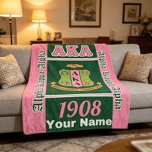 Op de afbeelding: Een roze en groene Alpha Kappa Alpha deken over een grijze bank. De deken heeft de letters "AKA" in roze, het jaar "1908" en de woorden "Your Name" in wit. De woorden "Alpha Kappa Alpha" staan verticaal op de zijkanten.