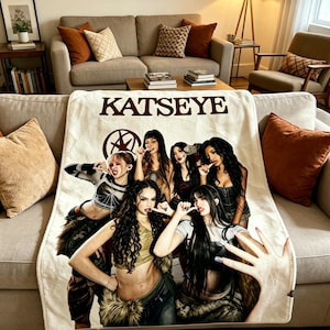Puede incluir: Una manta de forro polar color crema con el texto KATSEYE en letras marrón oscuro en la parte superior. La manta muestra una impresión fotográfica del grupo musical Katseye de seis miembros posando juntos, con una mano en primer plano.