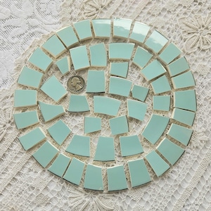 May include: A mosaic of light blue ceramic tiles arranged in a circular pattern. The tiles are various shapes and sizes, with a silver quarter coin in the center for scale. The tiles are on a white lace surface.