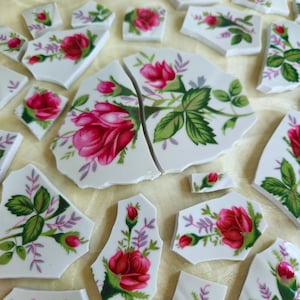 May include: A collection of white ceramic shards featuring a floral pattern of vibrant pink roses, green leaves, and small purple sprigs. The broken pieces vary in size and shape, displaying detailed botanical illustrations on a smooth surface.