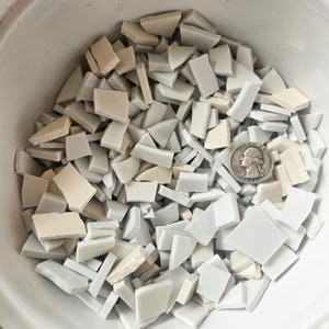 May include: A collection of broken ceramic tiles in shades of white and cream, with a silver quarter coin for size reference. The tiles are various shapes and sizes, suitable for mosaic art or craft projects.