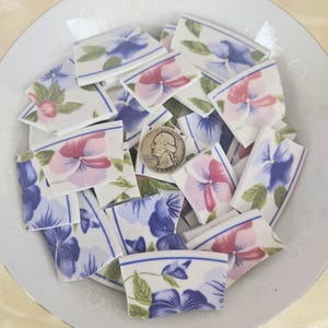 May include: A collection of broken ceramic pieces with floral designs in a white bowl. The pieces feature pink and purple pansies with green leaves. A quarter coin is included for size reference.