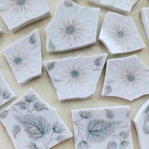 May include: White ceramic mosaic tiles with floral and leaf designs. The tiles are broken into irregular shapes, featuring embossed white flowers with tan centers and light blue accents. Some tiles have leaf patterns in gray and light blue.