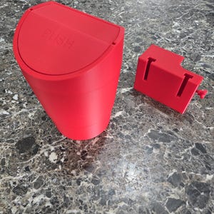 May include: A red, cylindrical waste bin with a curved lid. The lid is labeled "PUSH" in white text. A separate red component is positioned to the right. The bin is likely made of plastic and designed for small spaces.