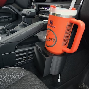 Pode incluir: Uma caneca Stanley laranja com tampa e alça brancas está em um porta-copos preto de carro. A caneca tem o texto "El Factory" e o logotipo da Stanley. Um canudo branco se estende da tampa. O porta-copos é montado em um carro.