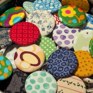 May include: A collection of colorful, fabric-covered buttons in various patterns and designs. The buttons feature floral, polka dot, geometric, and abstract prints in shades of red, green, blue, yellow, and purple. These buttons are ideal for sewing and craft projects.