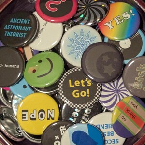 May include: A collection of colorful pinback buttons with various designs and text. Some buttons feature phrases like "YES!" and "Let's Go!", while others display images like a smiley face and a crescent moon with stars. The buttons are round and have a shiny finish.