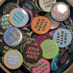 May include: A collection of round buttons with the message "STOP ASIAN HATE" in various colors and designs. Some buttons have the text in a circle, while others have the text in a straight line. The buttons are in a bowl.