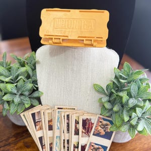 May include: A vintage Lipton Tea container in yellow, with the brand name printed on top. A collection of small, aged cards with various images are displayed in front of the container. Two small potted plants flank the display, set against a neutral backdrop.