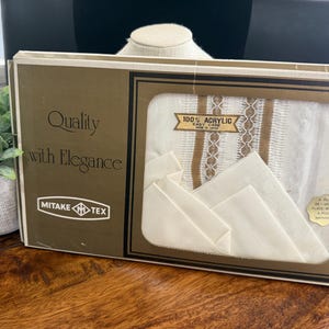 Puede incluir: Un juego de manteles individuales Mitake Tex en caja. La caja es marrón y dorada con las palabras "Quality with Elegance" impresas en la parte delantera. Los manteles individuales son blancos con detalles en marrón y dorado. La caja también tiene una etiqueta que dice "100% Acrylic Easy Care".
