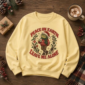 May include: A pale yellow crewneck sweatshirt featuring a central graphic of a frog wearing a Santa hat, a red patterned sweater, and a green scarf while holding a mug. The text reads PEACE ON EARTH LEAVE ME ALONE, surrounded by holly and ornaments.
