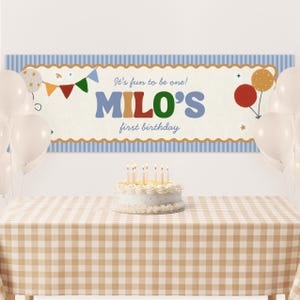 Może przedstawiać: Scena przyjęcia urodzinowego z banerem z napisem "It's fun to be one! MILO'S first birthday." Baner jest ozdobiony balonami i chorągiewkami. Ciasto ze świecami stoi na stole z kraciastym obrusem.