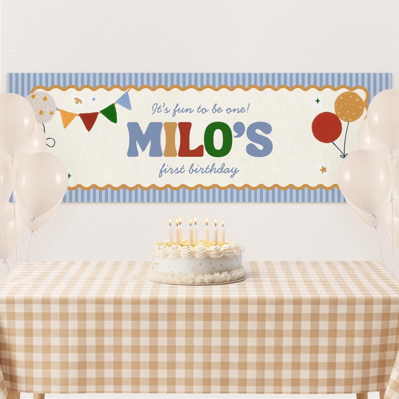 Może przedstawiać: Scena przyjęcia urodzinowego z banerem z napisem "It's fun to be one! MILO'S first birthday." Baner jest ozdobiony balonami i chorągiewkami. Ciasto ze świecami stoi na stole z kraciastym obrusem.
