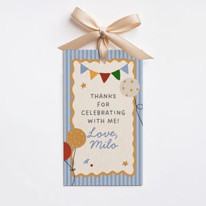 Może przedstawiać: Prostokątna etykieta prezentowa z jasnoniebieskim paskowanym tłem i falistą obw&oacute;dką. Etykieta zawiera tekst "THANKS FOR CELEBRATING WITH ME! Love, Milo" oraz ilustracje balon&oacute;w i proporczyk&oacute;w. U g&oacute;ry przymocowana jest beżowa wstążka.