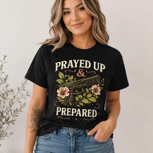 Peut inclure: T-shirt noir avec le texte "PRAYED UP & PREPARED" au-dessus et en dessous d'un dessin de pistolet entouré de fleurs roses et de feuilles vertes. Le modèle porte un jean bleu.