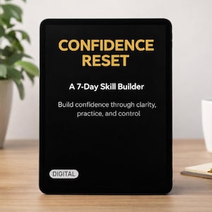 Op de afbeelding: Een zwarte tablet toont de tekst "CONFIDENCE RESET" in goud, met "A 7-Day Skill Builder" eronder. De tekst "Build confidence through clarity, practice, and control" is ook zichtbaar. Een witte mok, notitieblok en pen liggen op een houten oppervlak.